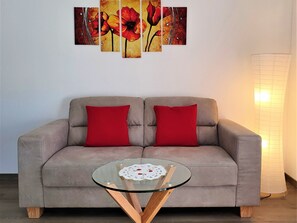A living room with a sofa, two red cushions, a glass coffee table, and a wall with a floral painting. - Ferienwohnungen Zur Heimat, (Grächen), by Interhome (Grächen)