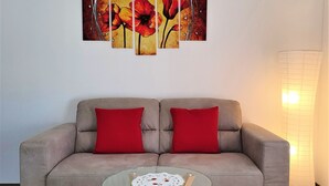 Un sofá gris con dos cojines rojos, una mesita de cristal con un mantel de encaje y una obra de arte floral colgada en la pared.