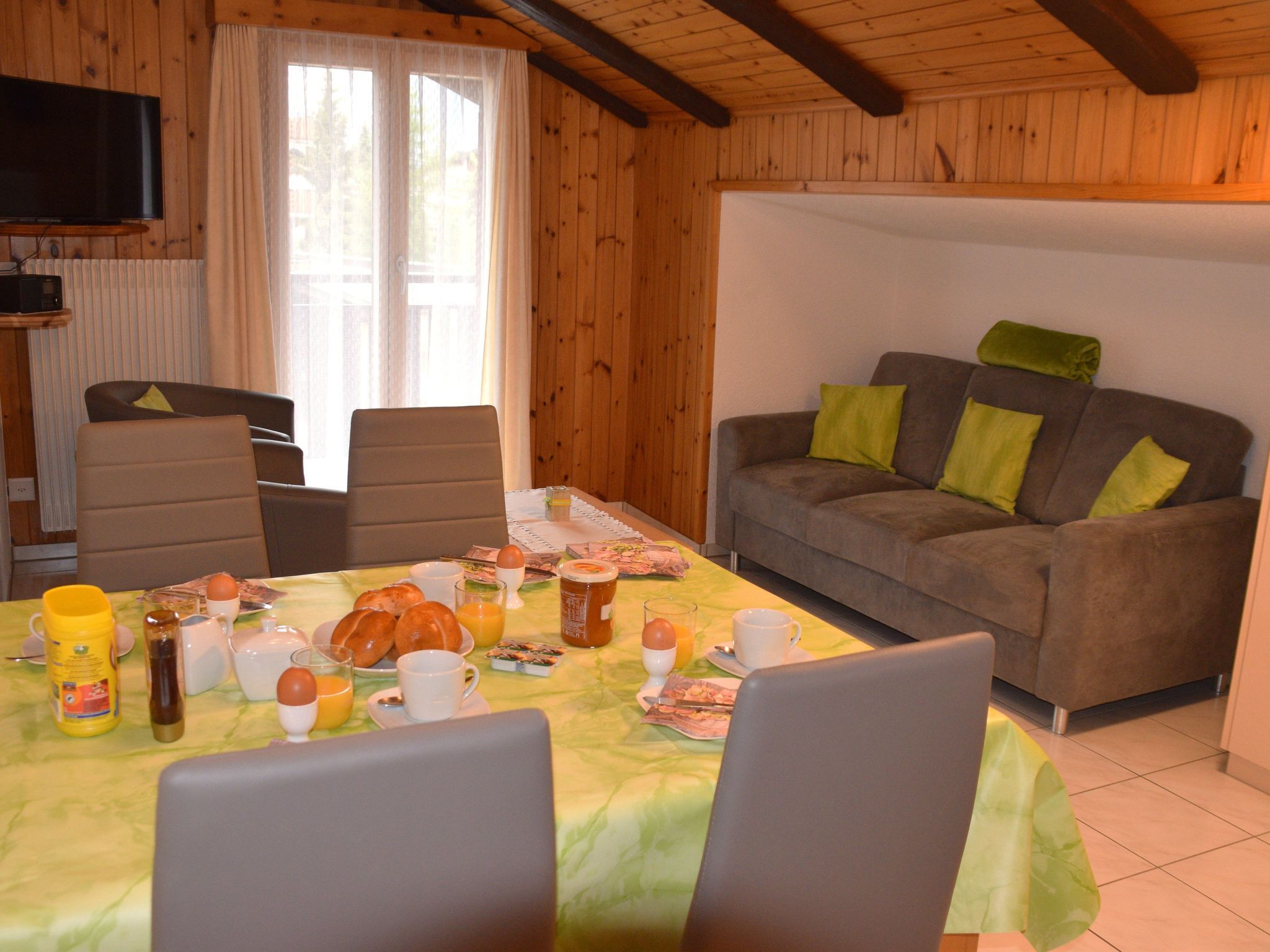 A cozy room with a dining table set for breakfast, a sofa with cushions, and a television mounted on the wall.