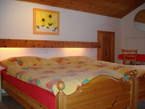 A bedroom with a wooden bed, floral bedding, a painting of sunflowers, a wooden ceiling, and a red chair. - Ferienwohnungen Zur Heimat, (Grächen), by Interhome (Grächen)