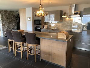 A modern kitchen with a central island, bar stools, a chandelier, and a stone wall. - Morion, Haus Alana, (Grächen), by Interhome (Grächen)