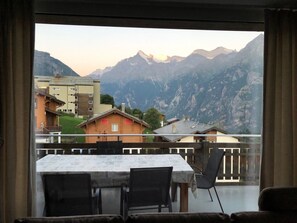 A room with a table and chairs, a view of mountains, and a balcony overlooking a body of water. - Morion, Haus Alana, (Grächen), by Interhome (Grächen)