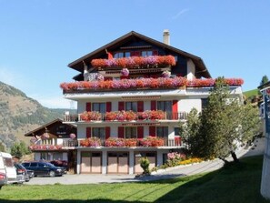 A multi-story hotel with balconies adorned with flowers, a mountain backdrop, and a clear sky. - Ferienwohnungen Schmiedstube 420, (Grächen), by Interhome (Grächen)