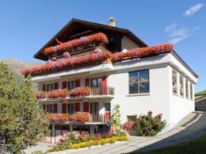 A multi-story building with balconies adorned with flowers, a clear sky, and a mountainous backdrop. - Ferienwohnungen Schmiedstube 420, (Grächen), by Interhome (Grächen)