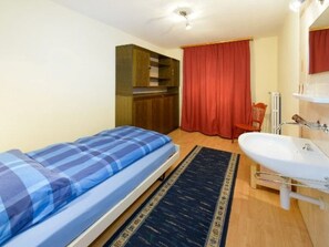 A small, single-bed room with a wooden wardrobe, a red curtain, a bathroom with a white sink, and a black rug. - Ferienwohnungen Schmiedstube 420, (Grächen), by Interhome (Grächen)