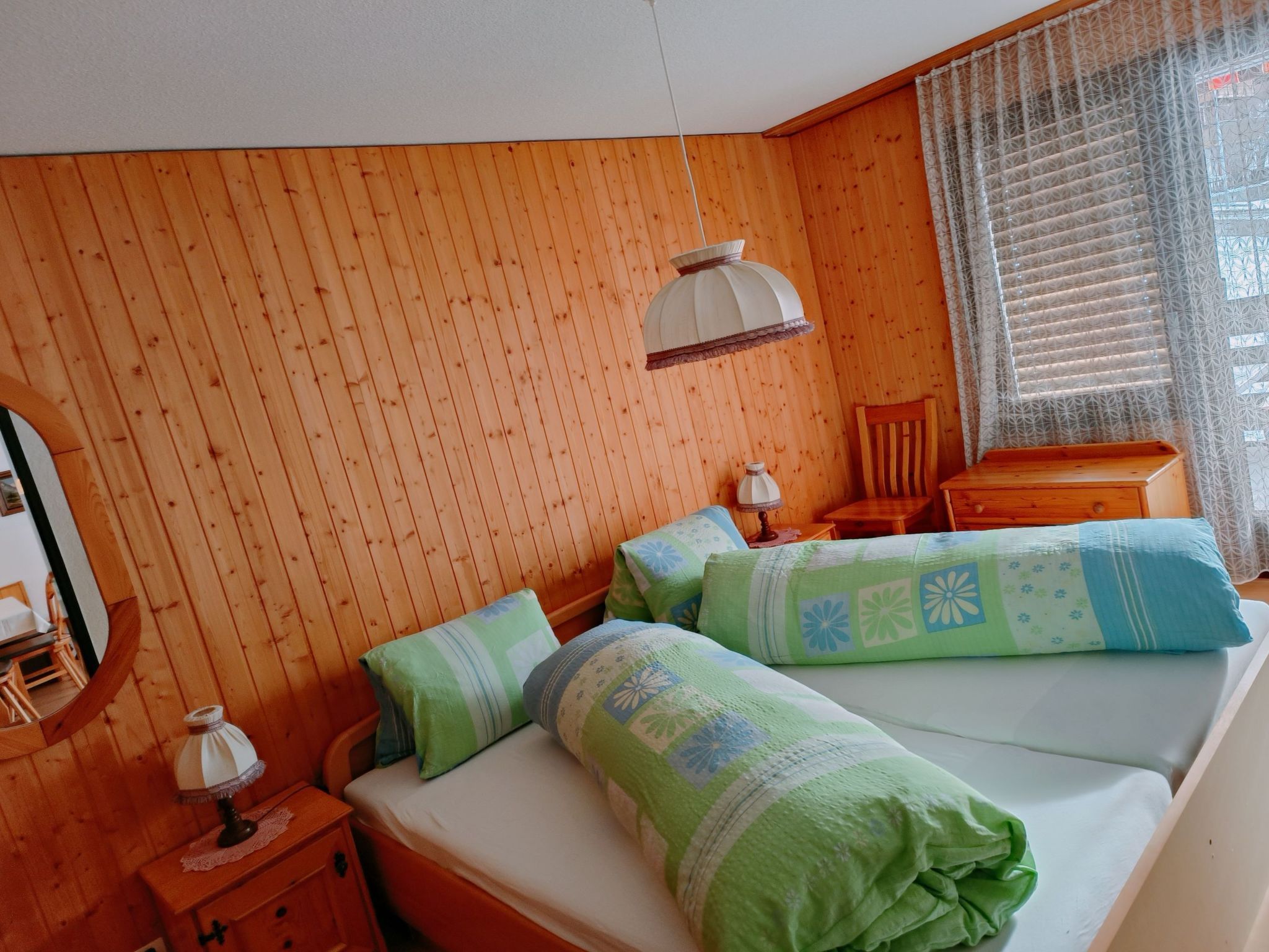 Ein Zimmer mit einer holzverkleideten Wand, einem Bett mit blaugrüner Bettwäsche, einem Nachttisch aus Holz mit einer Lampe, einem Sessel und einem Fenster mit gemusterten Vorhängen.