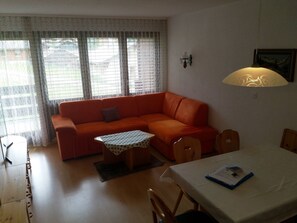 A living room with a sofa, a small table, a dining table, and a window with patterned curtains. - Nr.25,Haus Azurit, (Grächen), 1010 by Interhome (Grächen)