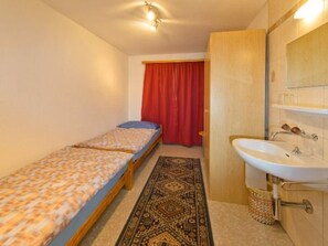 A small, single-bed room with a wooden bed, a red curtain, a white sink, and a patterned rug. - Ferienwohnungen Schmiedstube 420, (Grächen), by Interhome (Grächen)