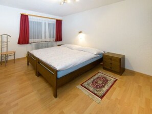 A bedroom with a wooden bed, a nightstand, a chair, a radiator, and a window with curtains. - Ferienwohnungen Schmiedstube 420, (Grächen), by Interhome (Grächen)