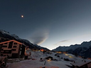Ein verschneites Bergdorf im Abenddämmerung mit beleuchteten Häusern und einem sichtbaren Mond.