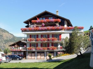 Ein mehrstöckiges Hotel mit Balkonen, die mit Blumen geschmückt sind, ein Parkplatz und eine Bergkulisse.