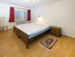 Ein Schlafzimmer mit einem hölzernen Bett, einem Nachttisch, einem Sessel, einem Fenster mit Vorhängen und einer Heizung.
