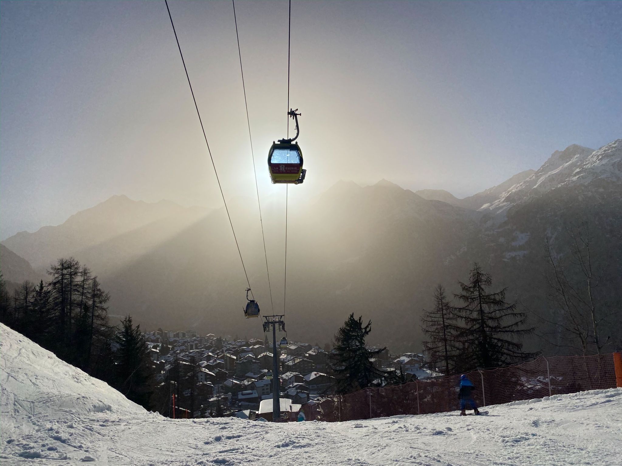 Una seggiovia con cabine sospese da cavi, con una montagna innevata sullo sfondo e una persona in piedi sulla pista.