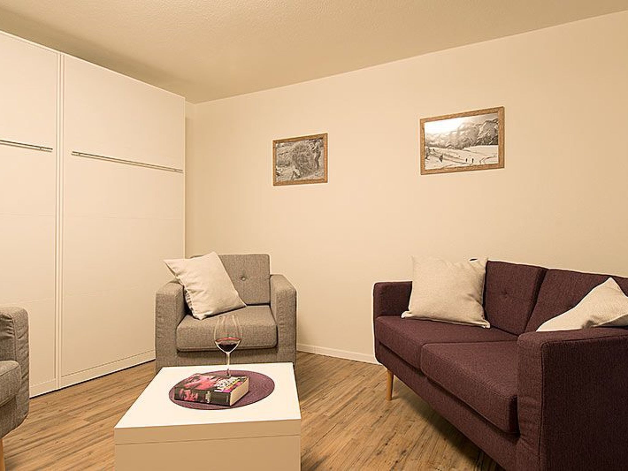 A living room with a grey sofa, a brown sofa, a coffee table with a book and a glass of wine, and two framed pictures on the wall.