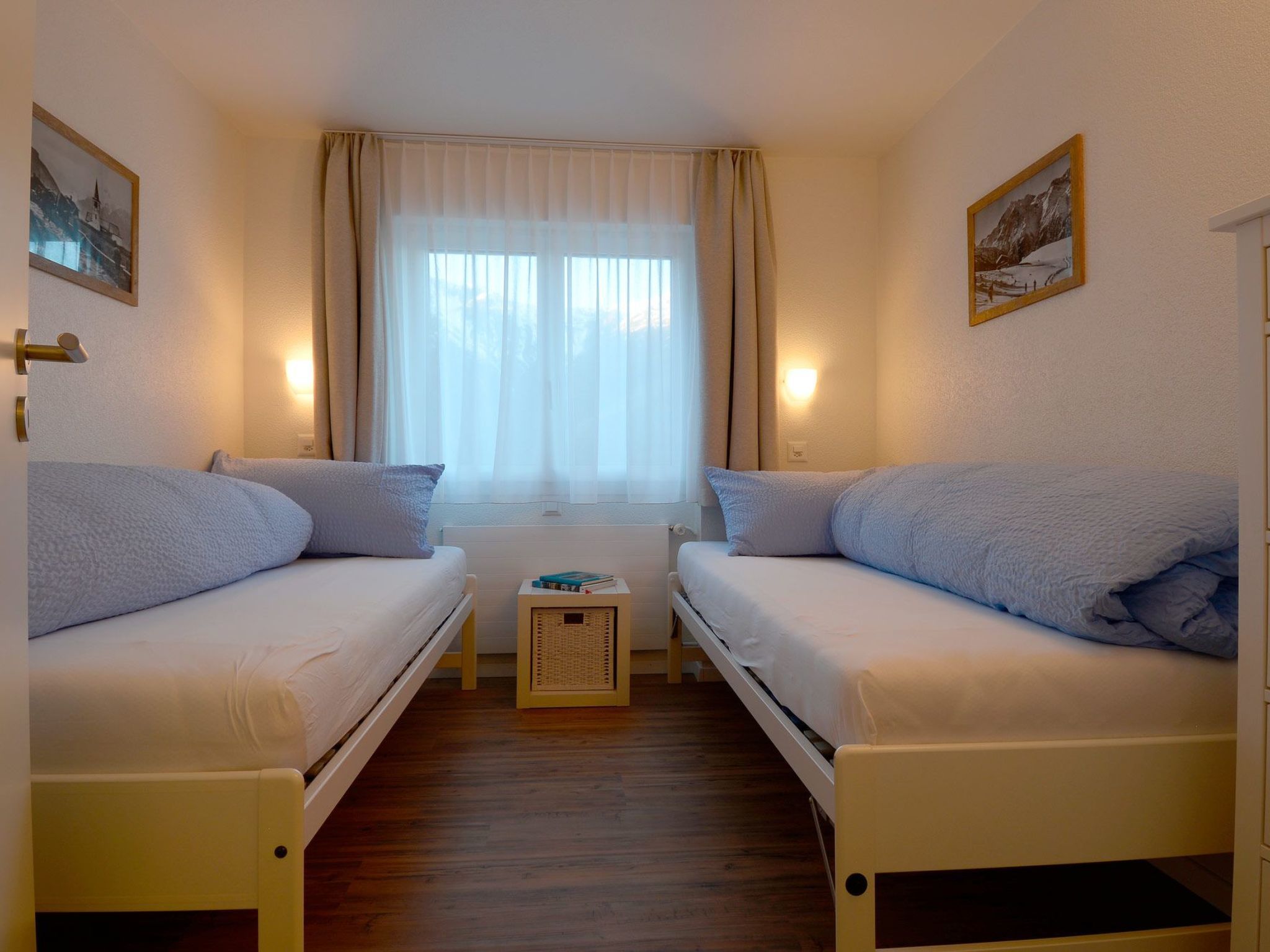 A small, cozy room with two single beds, a window with sheer curtains, and a radiator between the beds.