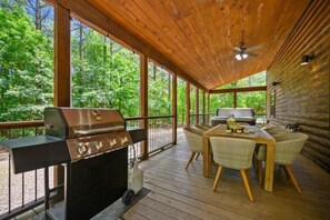 Una terraza exterior con una parrilla, mesa de comedor, sillas y vista a un bosque.