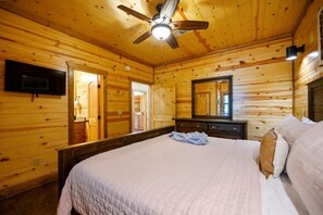 Un dormitorio tipo cabaña de madera con una cama, un ventilador de techo, un televisor montado en la pared y una puerta que da a otra habitación.