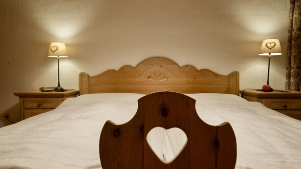 A bed with a wooden headboard featuring a heart cutout, flanked by two bedside tables with lamps, each lamp having a heart-shaped lampshade. - Goldblick, (Grächen), 1767+1034+2083 by Interhome (Grächen)