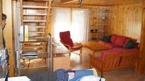 Een houten hut van binnen, met een houten bureau, boekenkasten, een rode stoel, een houten bank en een eettafel.