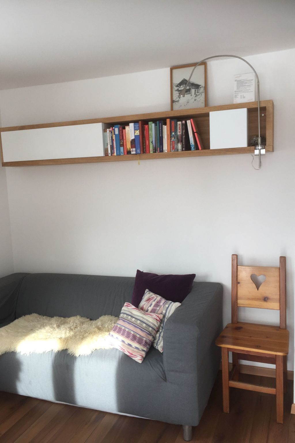 A grey sofa with a fluffy throw blanket and a patterned pillow, a wooden chair with a heart-shaped cutout, and a bookshelf with various books and a framed picture.