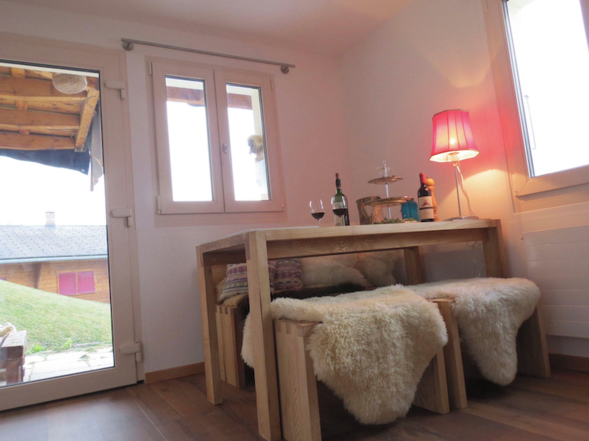 A room with a wooden table, two benches with fur throws, a lamp, and a window with a view of a building outside.