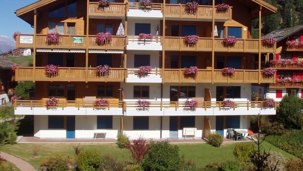 Un hôtel à plusieurs étages, doté de balcons ornés de fleurs, avec un ciel dégagé et un paysage montagneux en toile de fond.
