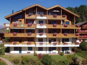 Ein mehrstöckiges Holzgebäude mit Balkonen, die mit Blumen geschmückt sind, unter blauem Himmel mit einer Bergkulisse im Hintergrund.
