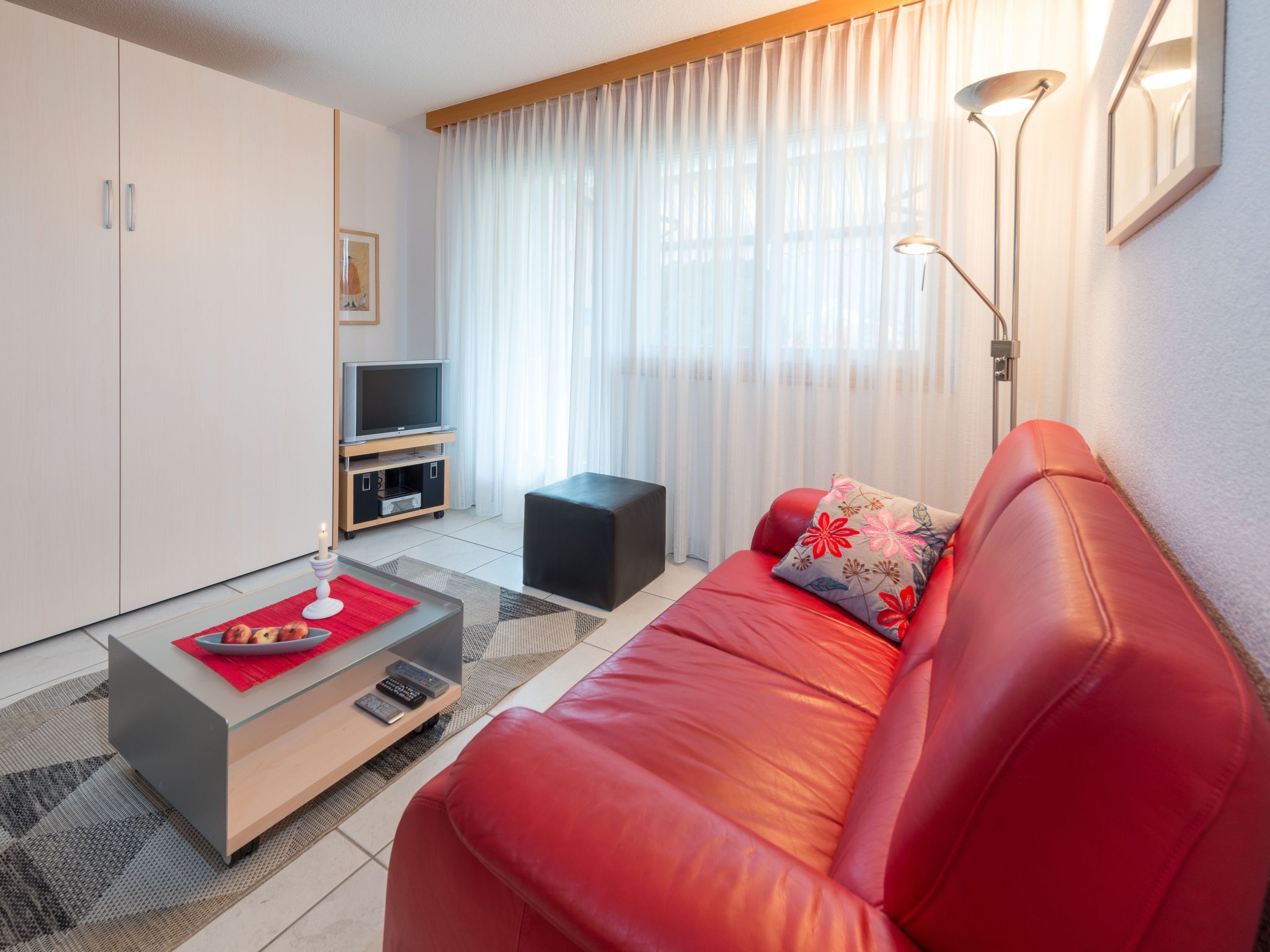 Een moderne woonkamer met een rode leren bank, een glazen salontafel, een flatscreen-tv op een houten dressoir en een groot raam met lichte gordijnen.