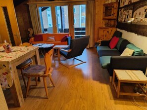 Ett mysigt vardagsrum med trägolv, ett matbord med stolar, en soffa och ett litet bord med en lampa.
