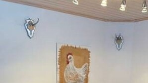 Un banc en bois avec une table, un tableau représentant un coq accroché au mur et une enseigne indiquant « Hetmaal ».