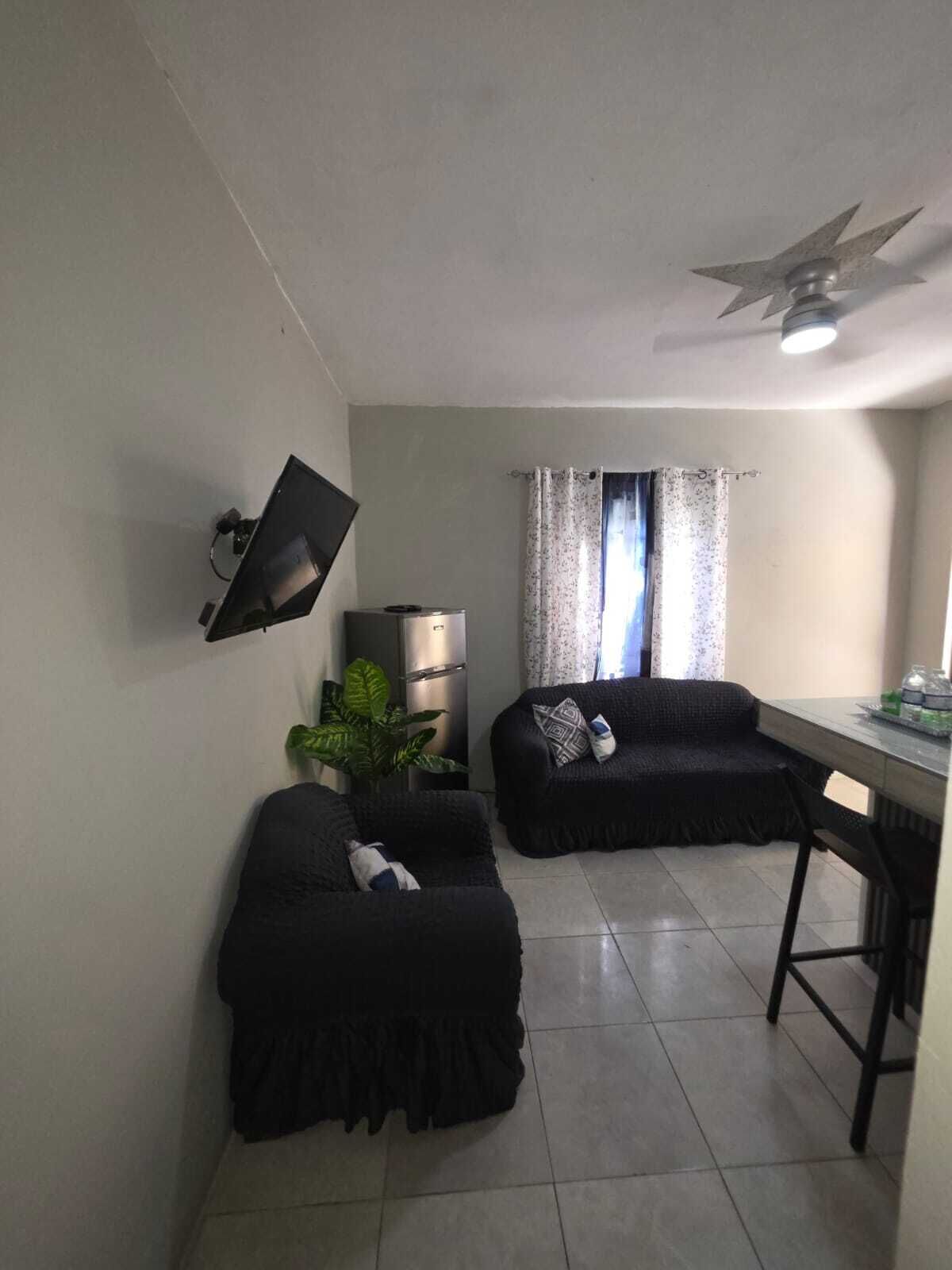Una sala pequeña con dos sofás negros, un televisor de pantalla plana montado en la pared, un ventilador de techo y una ventana con cortinas.