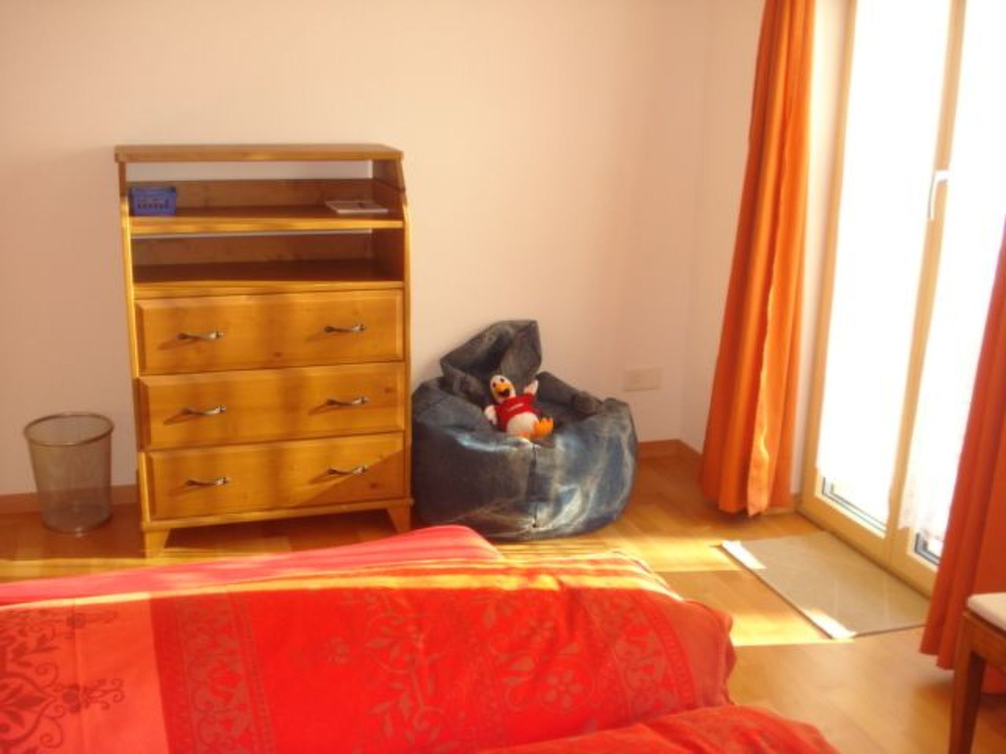 Een kamer met een houten dressoir, een loungestoel, een bed met een rode dekbedovertrek en een raam met gordijnen.