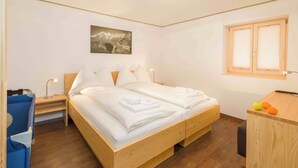 A bedroom with a wooden bed, a nightstand, a desk, and a window with blinds. - Lunic, AZapartments.ch, B&B, (Grächen), 1057 by Interhome (Grächen)
