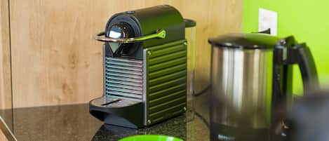 Une machine à café posée sur un plan de travail de cuisine, à côté d’un saladier vert et d’une bouilloire.