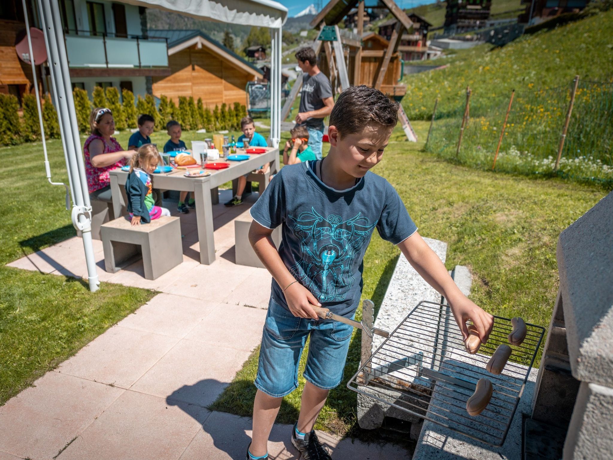 Een kind barbecuet worstjes tijdens een evenement buiten, met tafels en stoelen, op de achtergrond gebouwen en een grasheuvel.