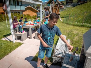 A child grilling sausages at an outdoor event with tables and chairs, set against a backdrop of buildings and a grassy hill. - Ferienwohnungen Lunic, AZapartments.ch, B&B, (Grächen), 1057 by Interhome (Grächen)