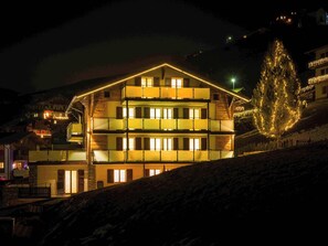 A multi-story building with balconies and illuminated windows, surrounded by a snowy landscape. - Lunic, AZapartments.ch, B&B, (Grächen), 1057 by Interhome (Grächen)