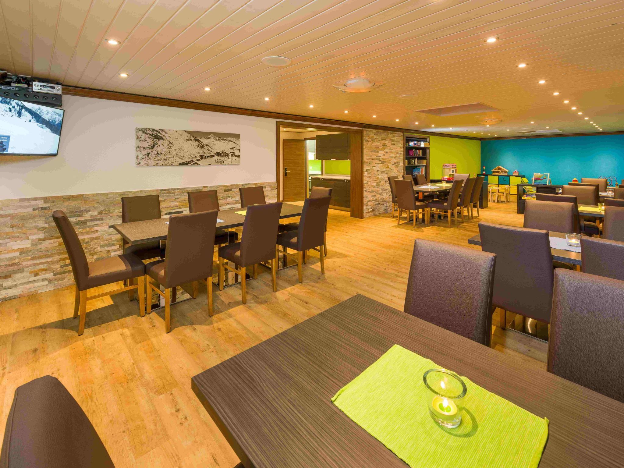 A restaurant with multiple dining tables and chairs, a television mounted on the wall, and a kitchen area visible through an open door.