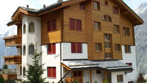 A multi-story wooden building with balconies, red shutters, and a mountain backdrop. - Thea, (Grächen), 1021 by Interhome (Grächen)