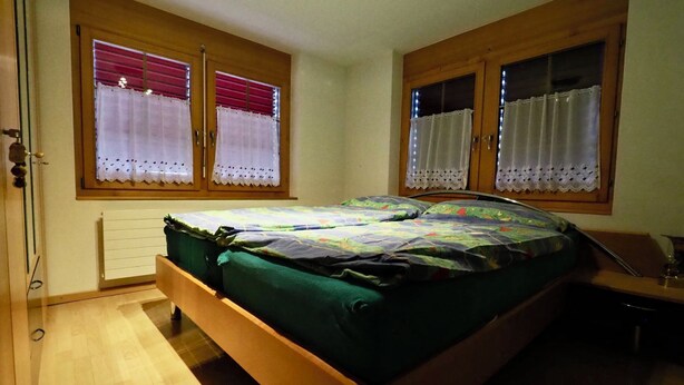 A bedroom with a wooden bed, two windows with curtains, a radiator, and a chair. - Thea, (Grächen), 1021 by Interhome (Grächen)