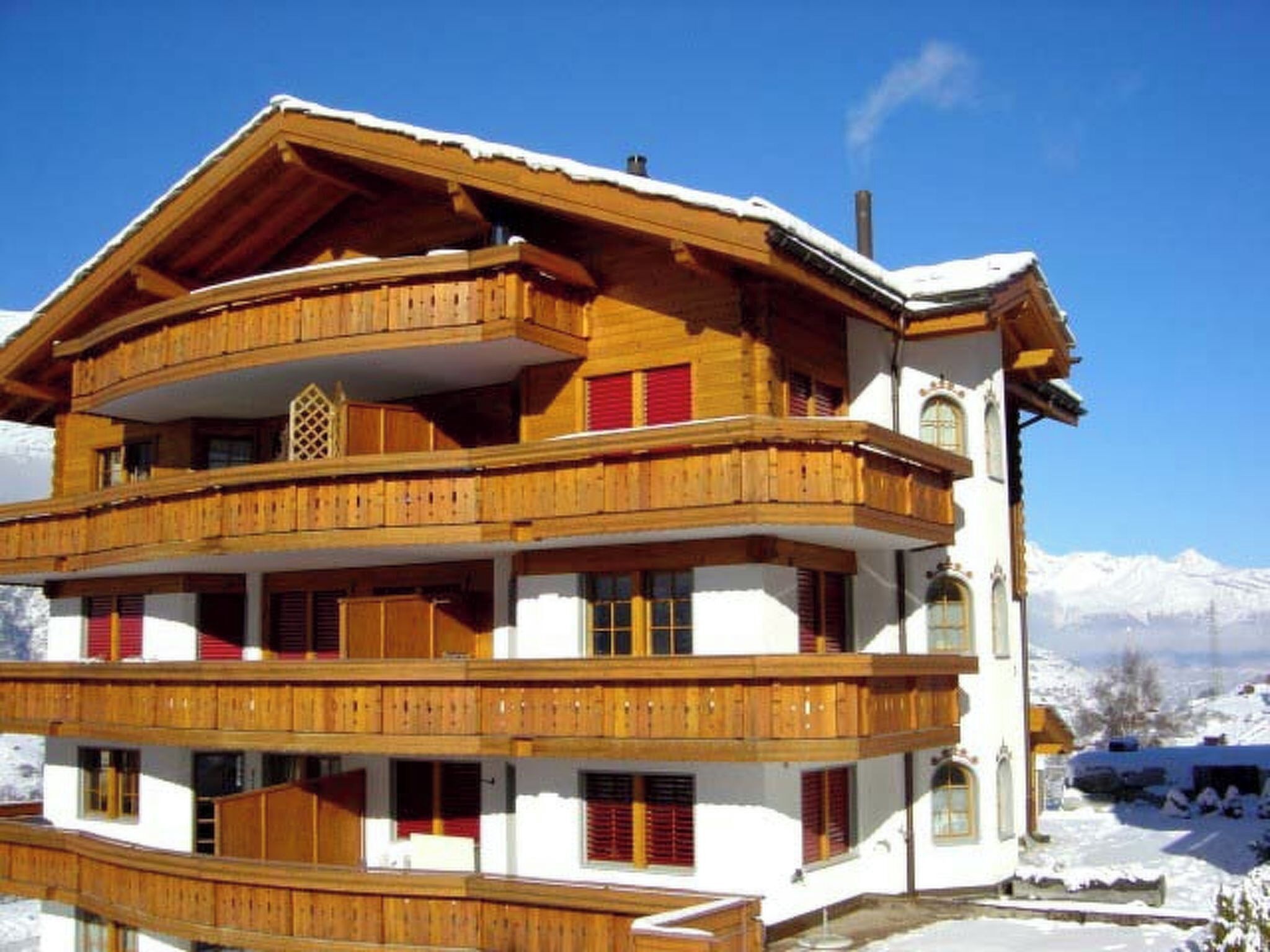 A multi-story wooden chalet with balconies, set against a snowy mountain backdrop.