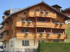 Un immeuble en bois à plusieurs étages, doté de balcons, dont certains sont ornés de jardinières fleuries, et sur lequel on peut lire l’inscription « Sunnegga ».