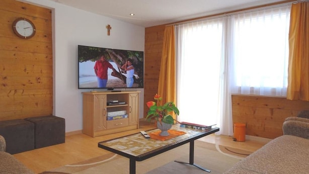 Un salon avec une télévision, des murs en bois, un crucifix, une fenêtre avec des rideaux et une table basse sur laquelle se trouvent des livres et une plante.