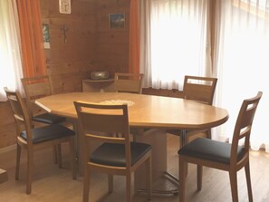 Ein runder Holztisch, umgeben von sechs Stühlen mit gepolsterten Sitzen, steht in einem Raum mit Holzwänden und einem Fenster mit durchscheinenden Vorhängen.