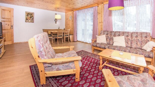 Ein Zimmer mit Holzwänden, einer Couch, Sesseln, einem Esstisch und einem Fernseher.