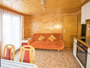 A cozy room with a wooden ceiling, a sofa with cushions, a small table with a remote control, and a kitchen area with cabinets and a countertop. - Ferienwohnungen Bergtreu, (Grächen), by Interhome (Grächen)