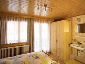 Makuuhuoneessa on sänky, ikkunassa verhot, patteri, puinen vaatekaappi sekä kylpyhuone, jossa on pesuallas ja peili.