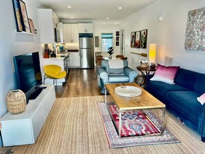 A modern living room with a TV, sofa, coffee table, and a kitchen in the background. - The Little Retreat Unit A - Walk to Shops & Beach (Cape Charles)