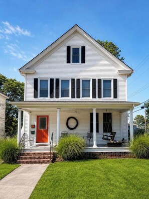A two-story white house with a red door, a porch with chairs, and a wreath. - The Little Retreat Unit A - Walk to Shops & Beach (Cape Charles)
