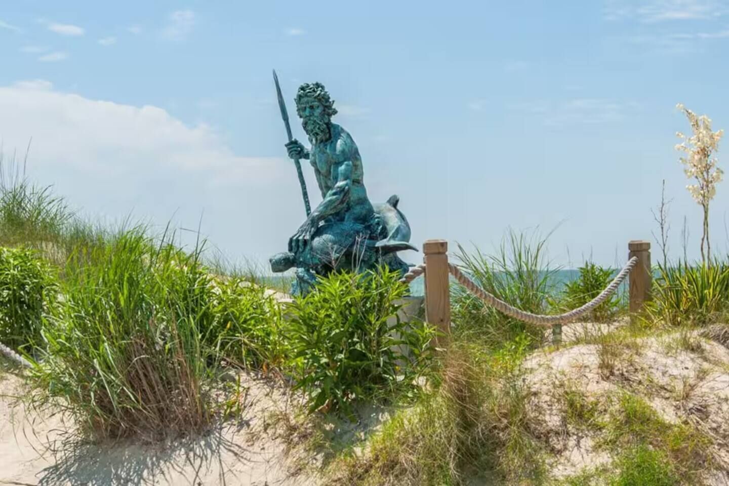 A statue of a figure holding a spear, surrounded by sand dunes and vegetation.